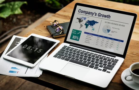 A laptop on a desk showing a business growth dashboard beside a tablet and printed reports